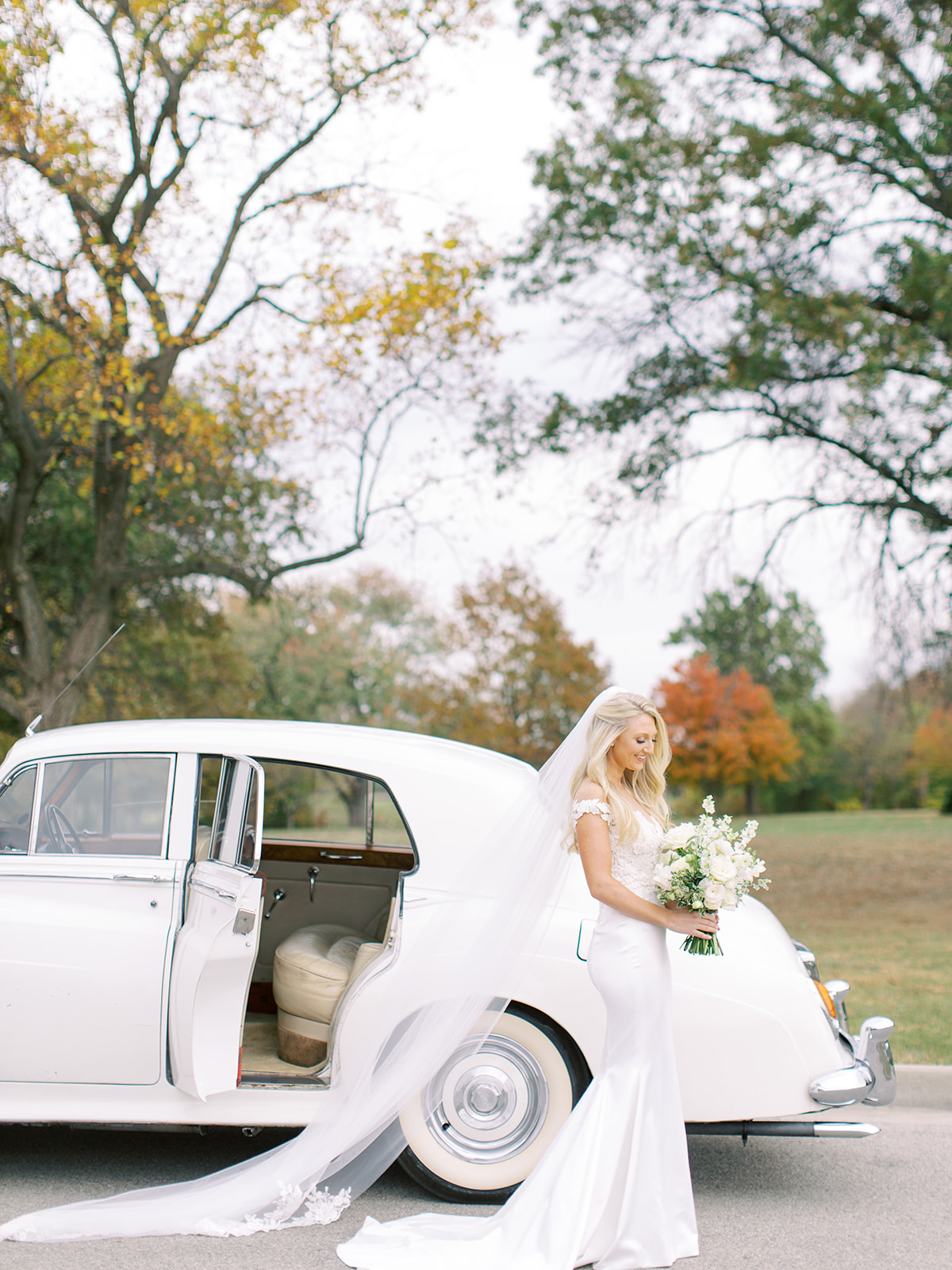 Bride smiling at bouquet in front of classic car