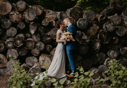 Bride and groom post together in front of outdoorsy background of logs and greenery.